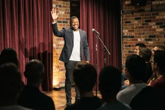 Humoriste noir masculin sur scène dans un comedy club intimiste avec micro et rideau bordeaux